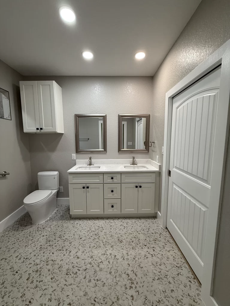 Modern bathroom featuring a double sink vanity with two mirrors, a toilet, and beige walls. The floor is tiled with a speckled pattern, and there is a cabinet above the sink.