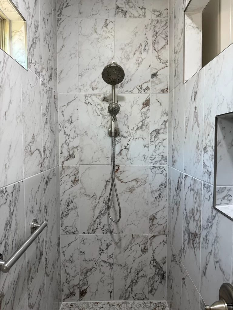 A modern shower featuring white marble tiles with gray veining, a wall-mounted showerhead, and a silver grab bar.