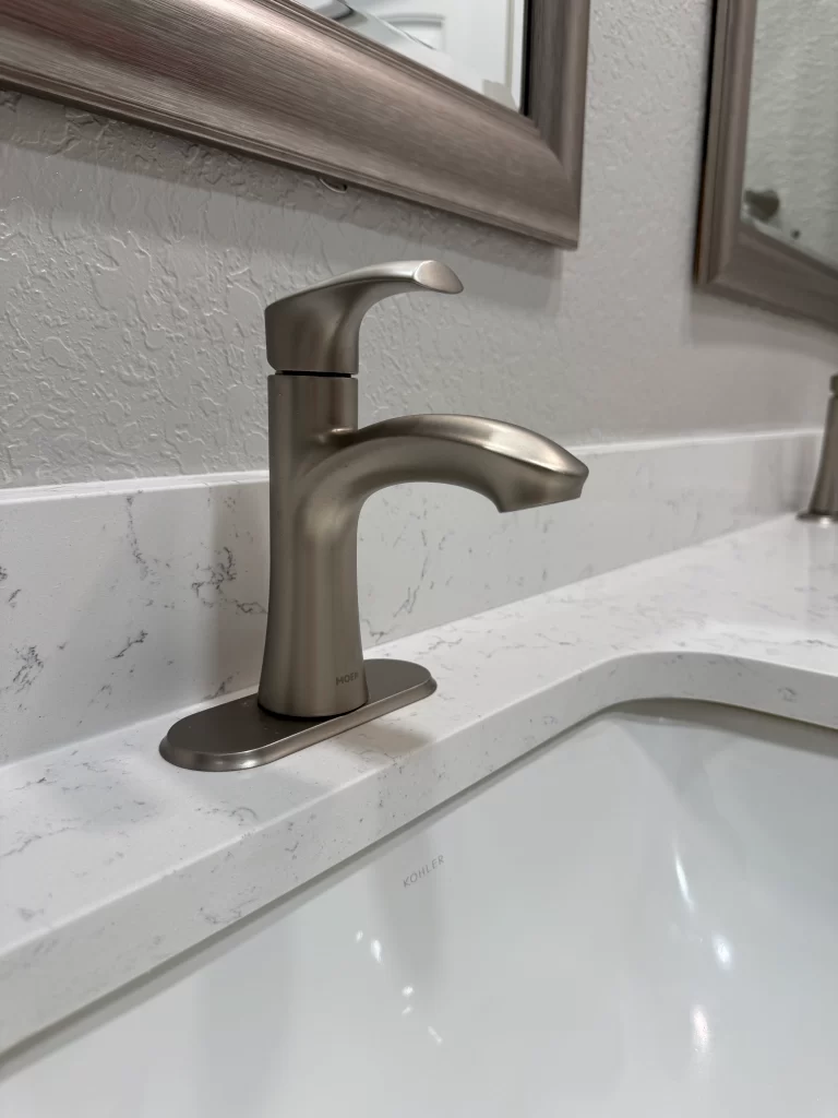 Close-up of a modern satin nickel faucet installed on a white countertop with a rectangular sink and a mirror in the background.