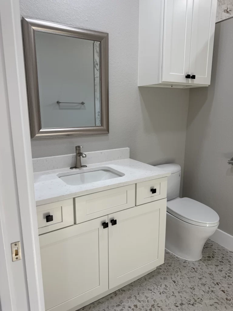 A modern bathroom featuring a white vanity with black handles, a sink, and a large mirror above it. A toilet is positioned beside the vanity, with gray walls and a pebble floor.