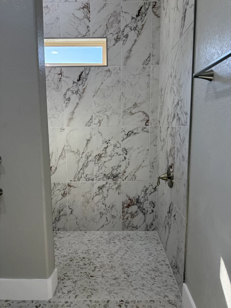Interior view of a modern shower featuring white marble tiles on the walls, a small window allowing natural light, and a pebble stone floor.