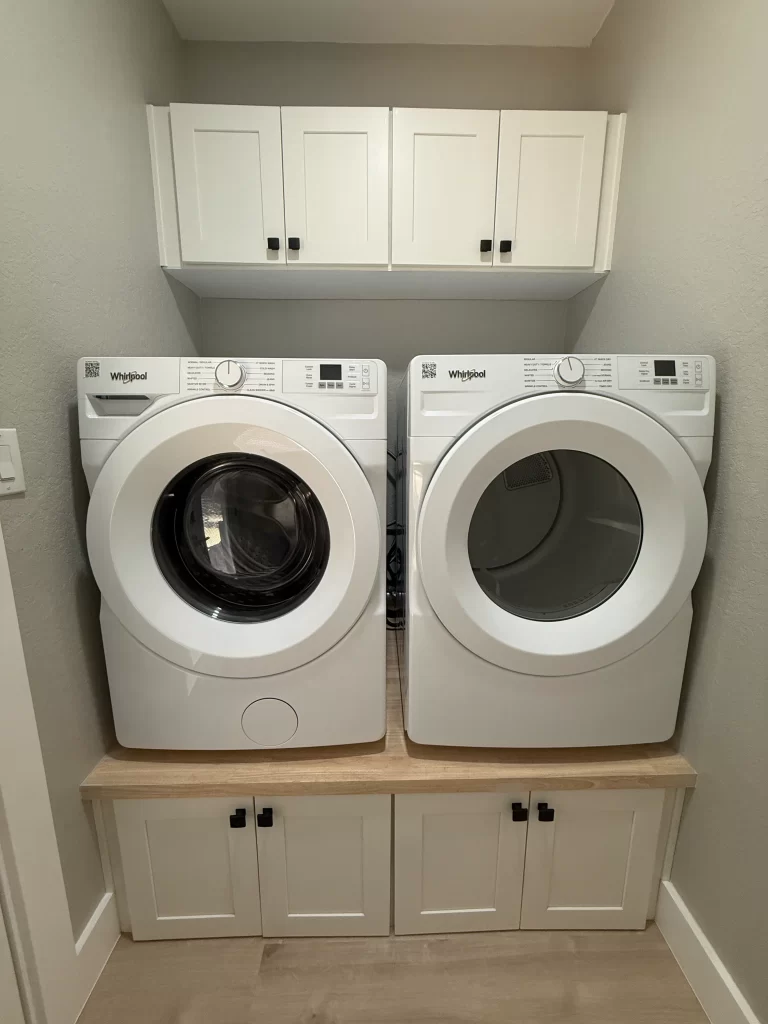A pair of white Whirlpool washing machine and dryer side by side in a laundry area, with white cabinets above and a wooden countertop below.