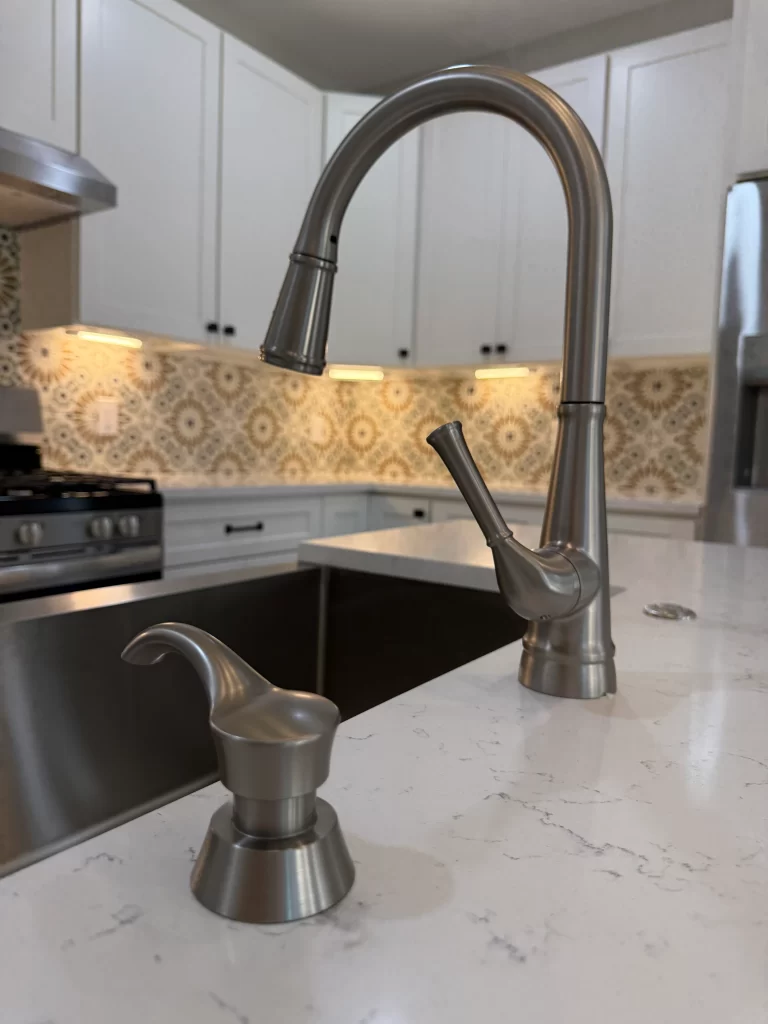 Close-up of a modern kitchen faucet and soap dispenser on a marble countertop, with white cabinets and decorative backsplash in the background.