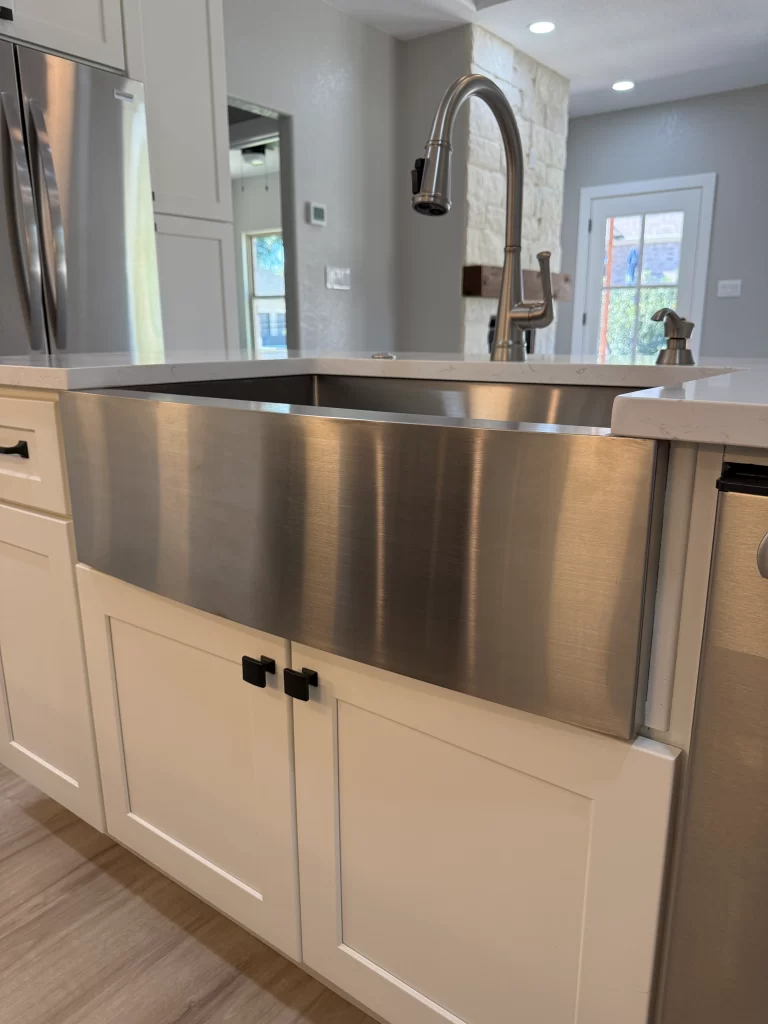 A close-up view of a stainless steel farmhouse sink integrated into white cabinetry, featuring black handles. The kitchen has modern appliances and a neutral color palette.