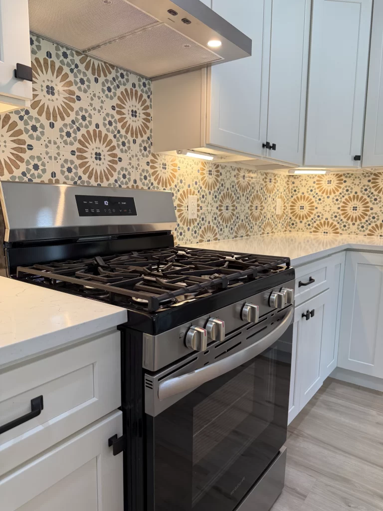 A modern kitchen featuring a stainless steel gas range with a black finish, white cabinetry, and an intricately patterned tile backsplash in earthy tones.