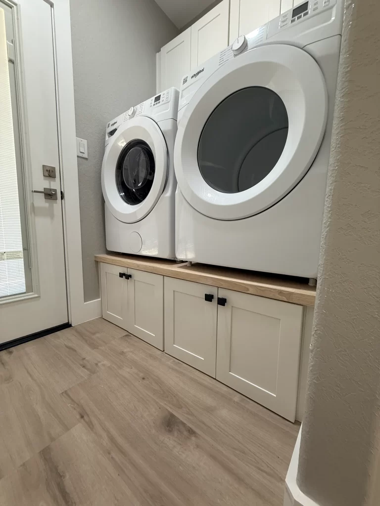 A modern laundry room featuring stacked white washer and dryer units next to a door, with a wooden shelf beneath them and white cabinetry below.