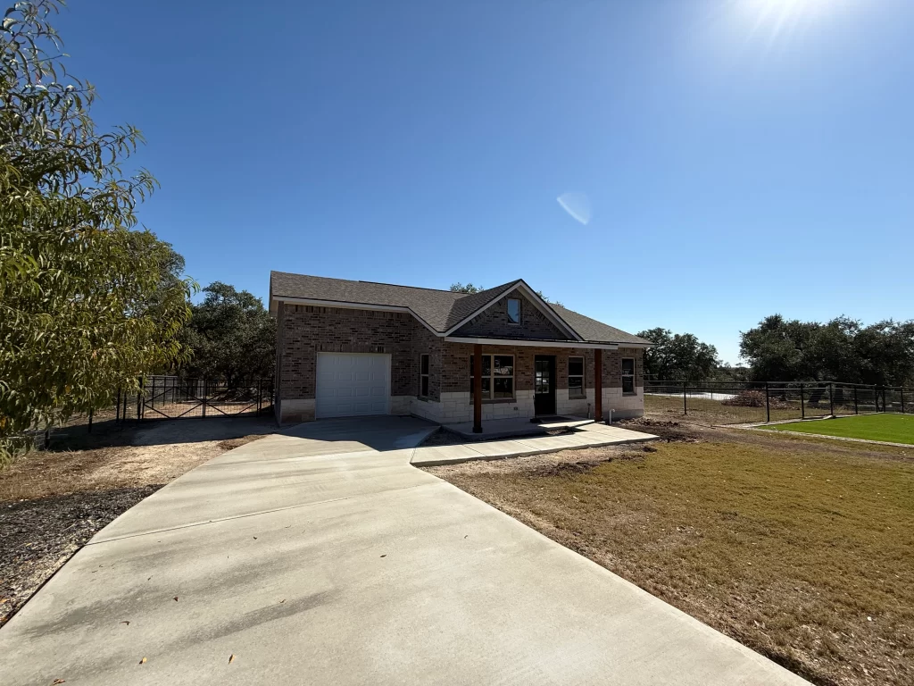 A single-story brick house with a front porch, surrounded by grass and trees under a clear blue sky.