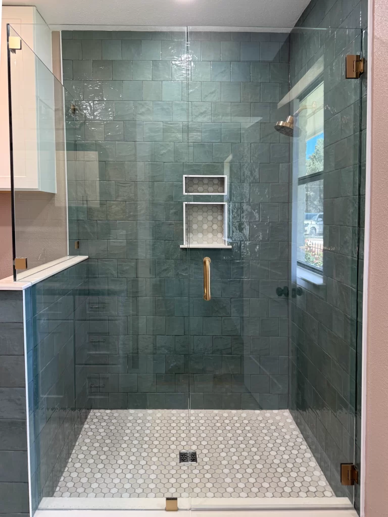 Modern glass shower with green subway tile walls and a white hexagonal tile floor, featuring a built-in shelf, brass fixtures, and natural light coming through a nearby window.