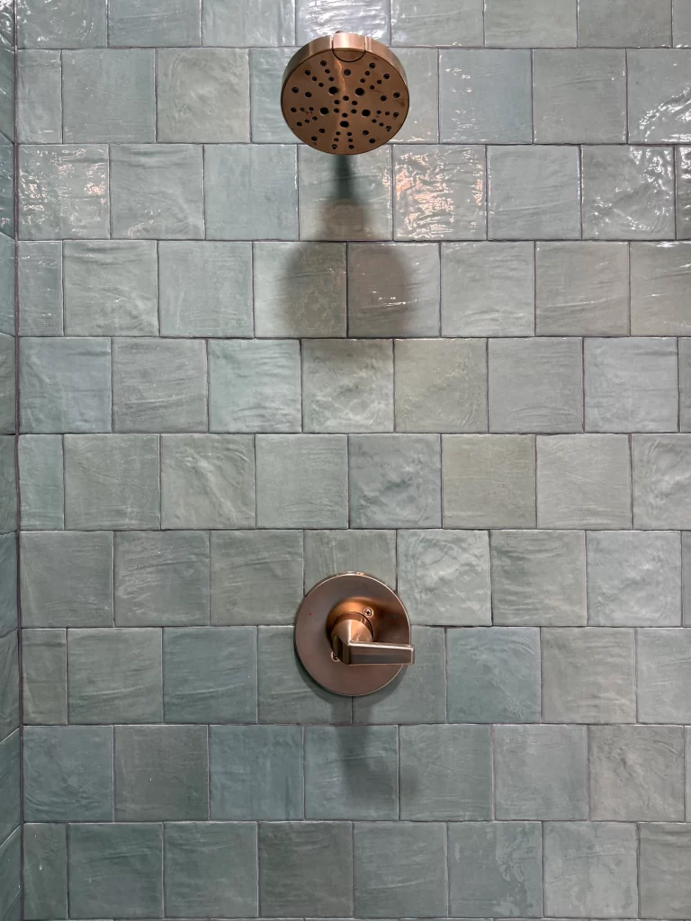 Close-up of a shower head and water control handle mounted on a textured blue-green tile wall.