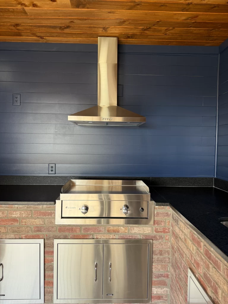 Outdoor kitchen featuring a stainless steel range hood and a built-in grill on a brick countertop with cabinets below.