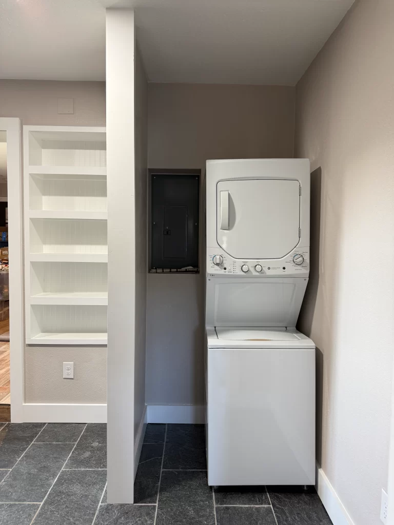 A compact laundry area featuring a stacked washer and dryer next to a wall with a black electrical panel and a white shelf unit in the background.