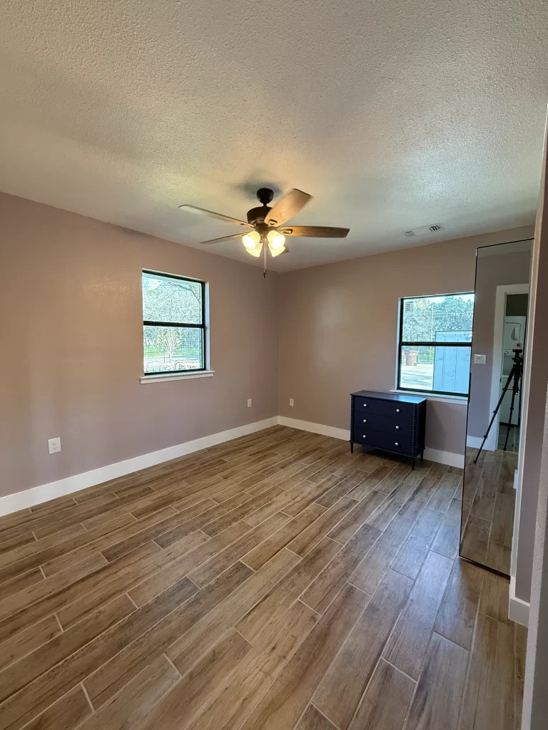 A spacious room with light purple walls, wooden tile flooring, and a ceiling fan. Features two windows for natural light and a small dresser against the wall.