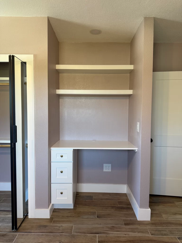 A light-colored room featuring a small desk with three drawers, two shelves above, and a mirrored closet door on the left.