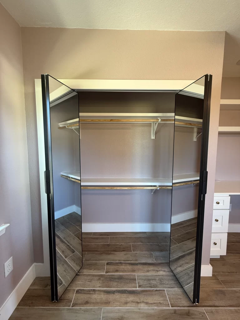 A walk-in closet with mirrored double doors, showcasing wooden shelving and a light-colored interior.