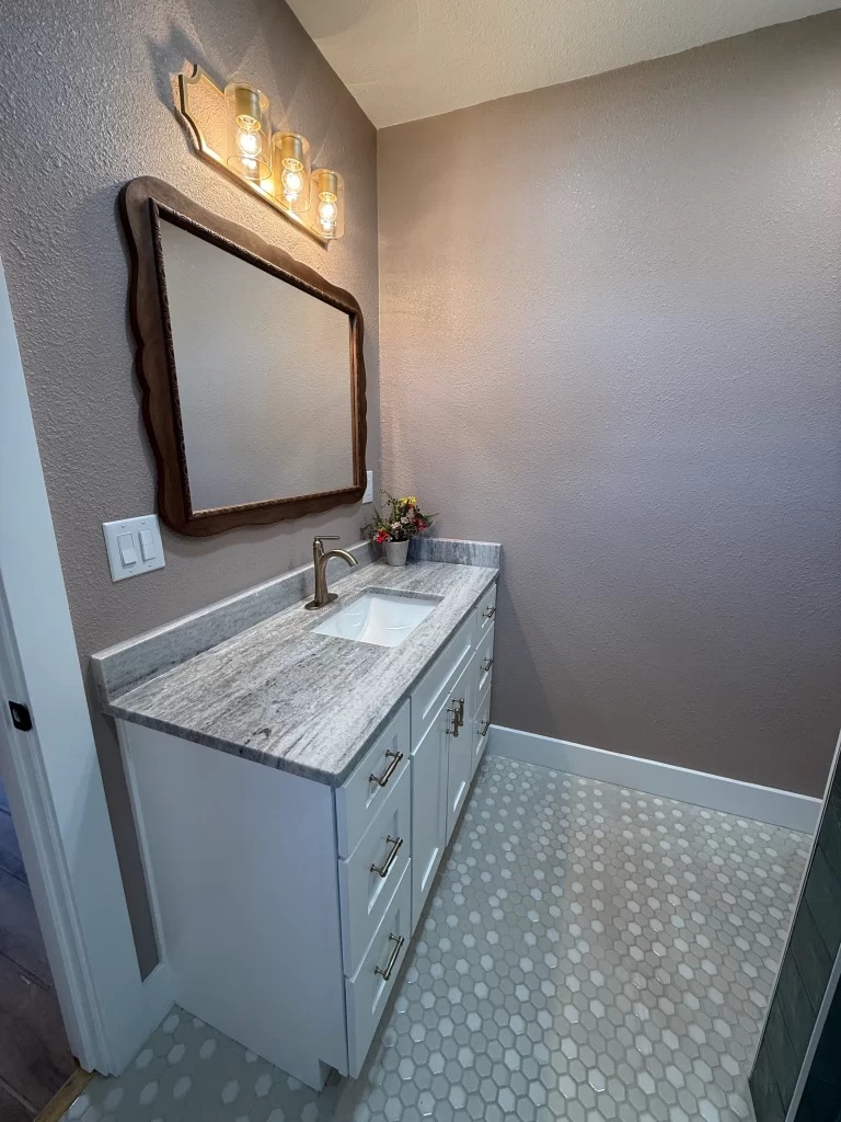 A modern bathroom featuring a white vanity with a gray countertop, a decorative mirror, and stylish lighting above. The floor is covered in hexagonal tiles, and a small vase of flowers adds a touch of decor.