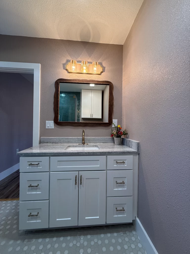 Modern bathroom vanity with white cabinetry, a grey countertop, and a large mirror. The space features a light fixture above the mirror and a small vase of flowers.