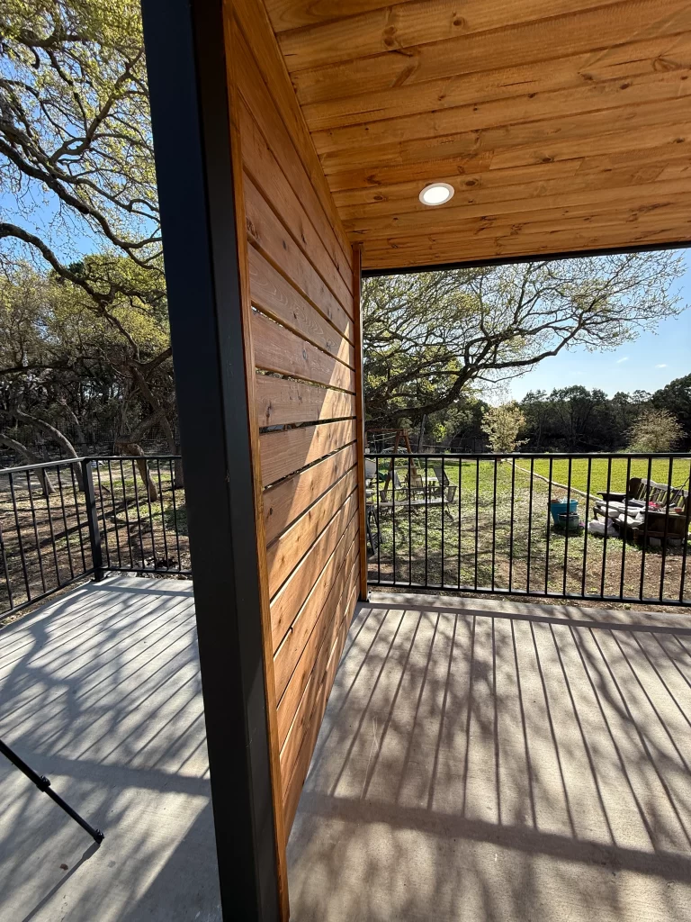 View from a wooden porch featuring a wall of horizontal wooden planks, with a ceiling light and a railing overlooking a green field and trees.