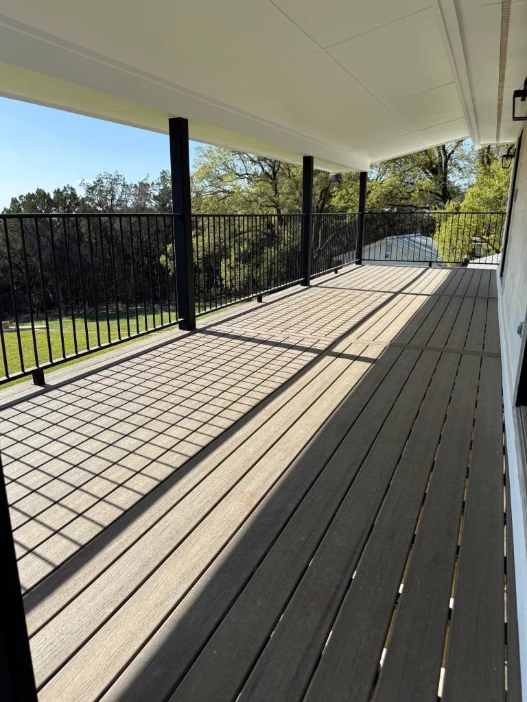 A spacious wooden deck with metal railing, under a covered porch, displaying sunlight and shadows on the surface.