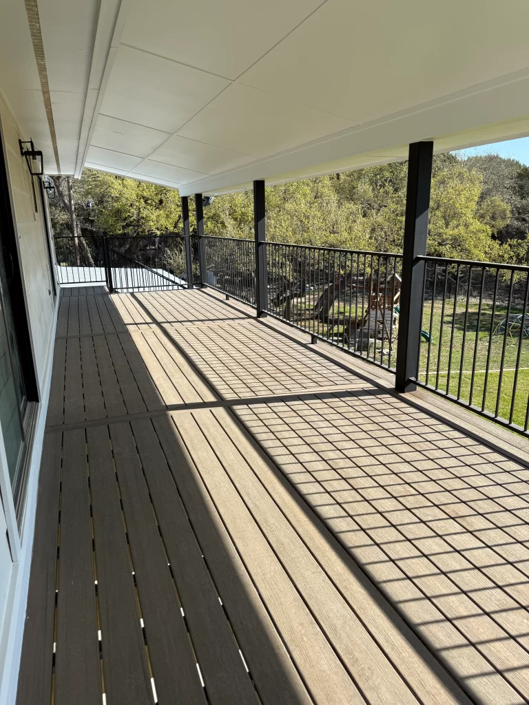 A well-lit deck area featuring wooden flooring and a black railing, surrounded by greenery.