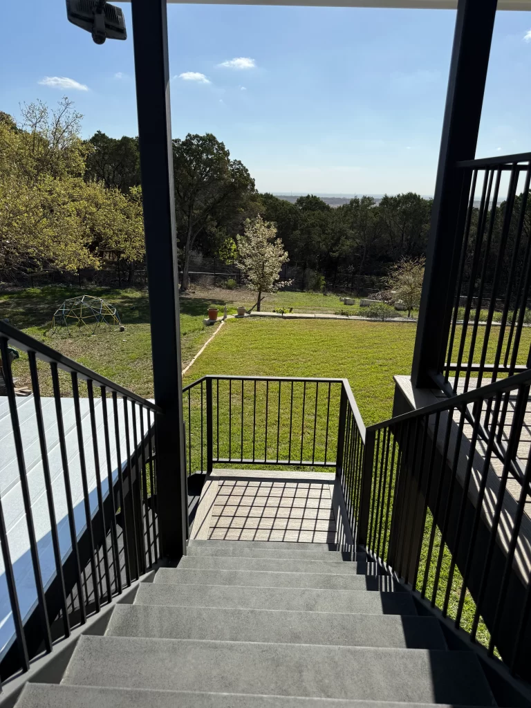 View from a staircase leading down to a grassy yard surrounded by trees on a sunny day.