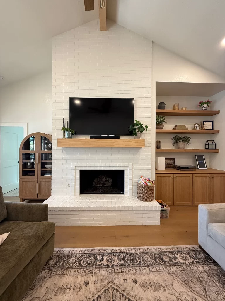 A modern living room featuring a white brick fireplace, mounted flat-screen TV, and wooden shelves with decor. A dark green sofa and a light gray couch are positioned in front of the fireplace, with a patterned rug covering the floor.