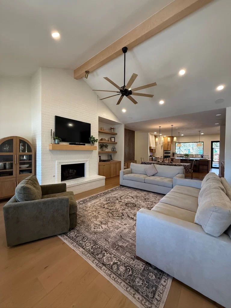 Spacious living room featuring a beige sectional sofa, two green armchairs, a decorative rug, a white brick fireplace, and an entertainment system mounted on the wall.