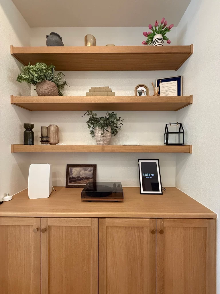 A wooden shelf display with decorative items including plants, books, and a framed picture, above a wooden cabinet with a clock and a record player on top.