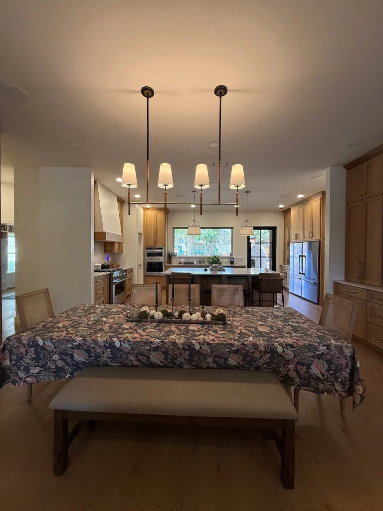 A modern kitchen with a dining table covered by a floral tablecloth, surrounded by a bench and chairs, under overhead pendant lighting. The kitchen features wooden cabinets and appliances in the background, with natural light coming through a window.
