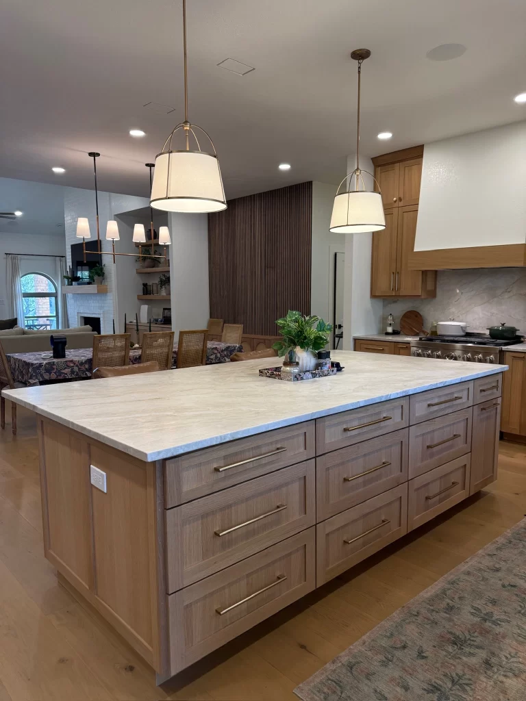 A modern kitchen featuring a large wooden island with several drawers and a light-colored countertop, surrounded by stylish pendant lights. In the background, there are dining chairs and plants, creating an inviting atmosphere.