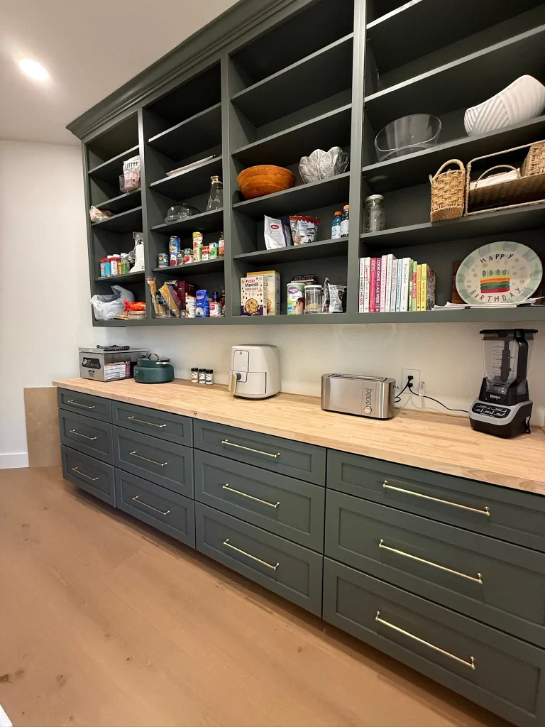 A modern kitchen storage area featuring green cabinetry with golden handles, open shelves holding various kitchen items, and a wooden countertop. Appliances like a blender, toaster, and air fryer are neatly arranged on the counter.