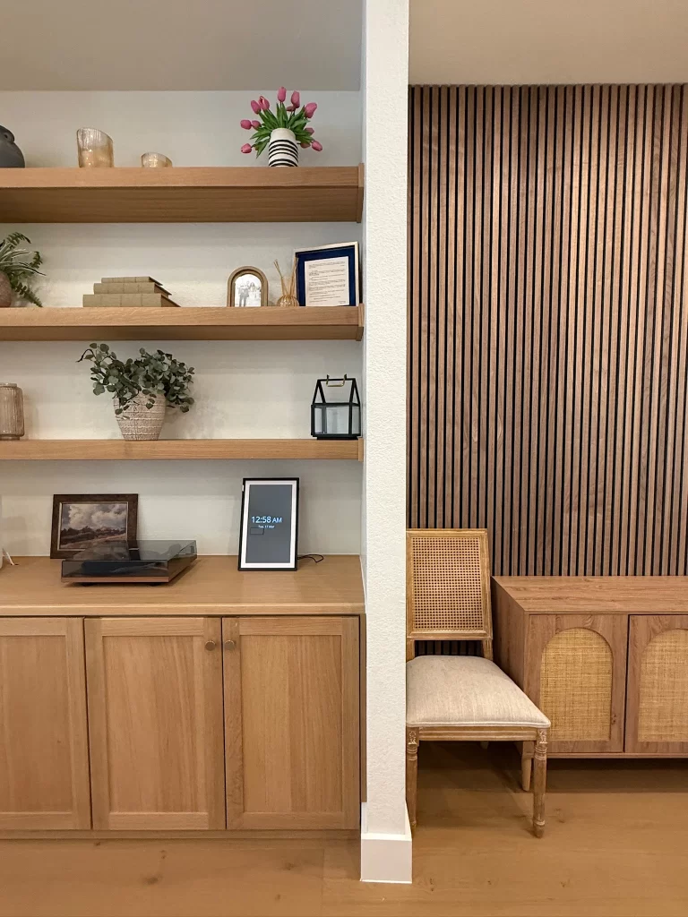 A minimalist interior scene featuring wooden shelves with decorative items, including plants, books, and a framed picture. A digital clock displays the time on a nearby cabinet, alongside a light-colored chair and a wooden sideboard with woven elements.