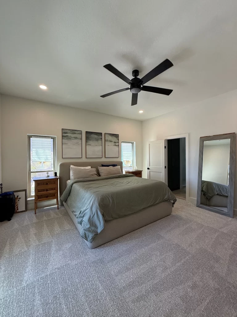 A modern bedroom featuring a large bed with a green blanket, two pillows, a ceiling fan, and artwork on the wall. Natural light coming through windows and a full-length mirror nearby.