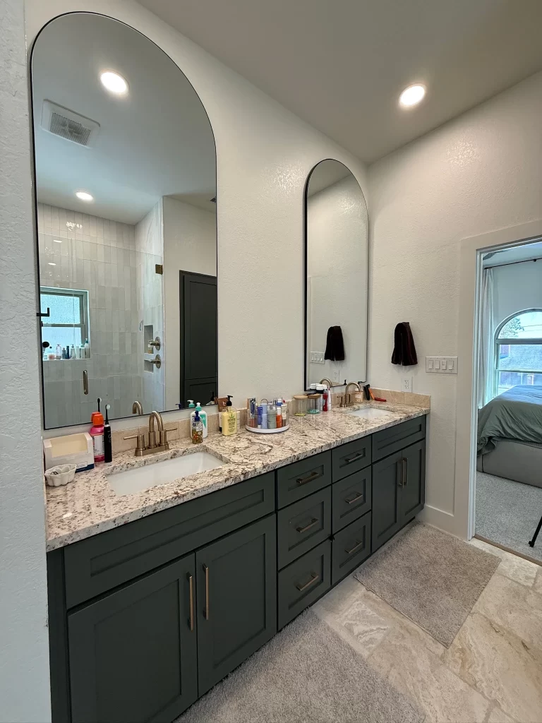 Modern bathroom featuring a double sink vanity with dark cabinetry, a granite countertop, and large round mirrors. Also includes a glass shower and an adjacent doorway leading to a bedroom.