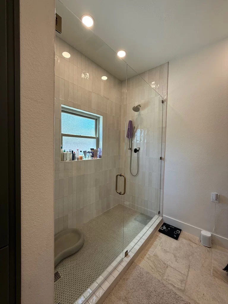 A modern shower with glass doors, featuring a tiled interior, a showerhead, and a small window allowing natural light. Various bottles of toiletries are visible on the ledge.