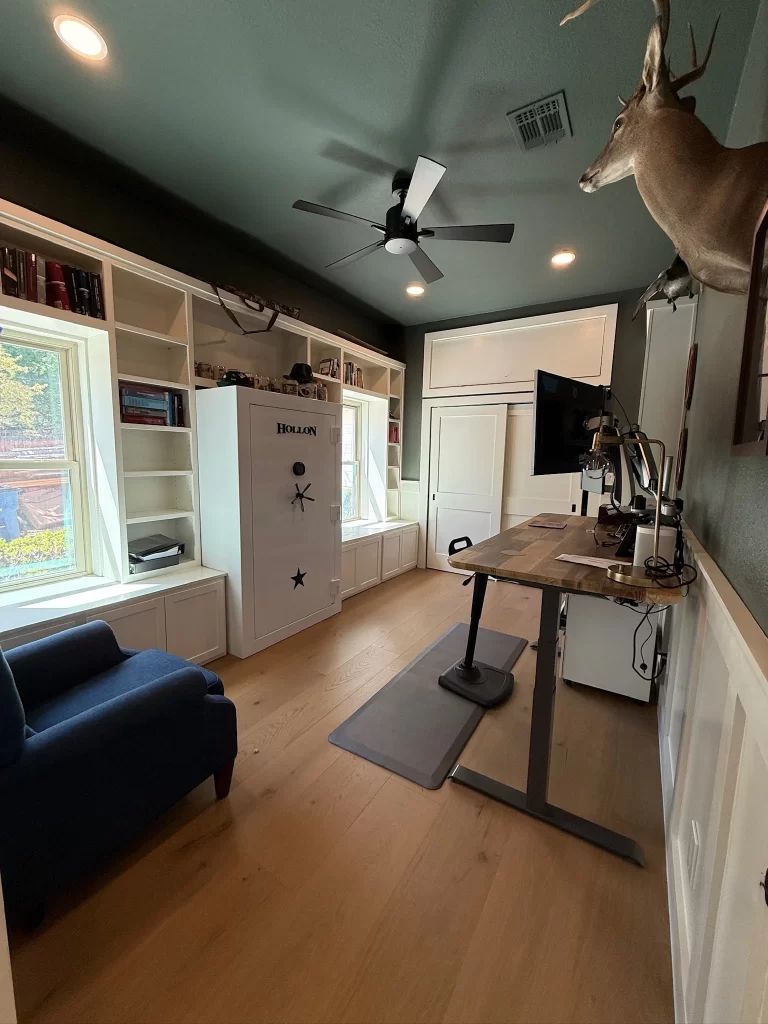 A modern home office featuring a standing desk, a blue accent chair, built-in bookshelves, and a deer head mounted on the wall. Bright lighting and a ceiling fan enhance the workspace ambiance.