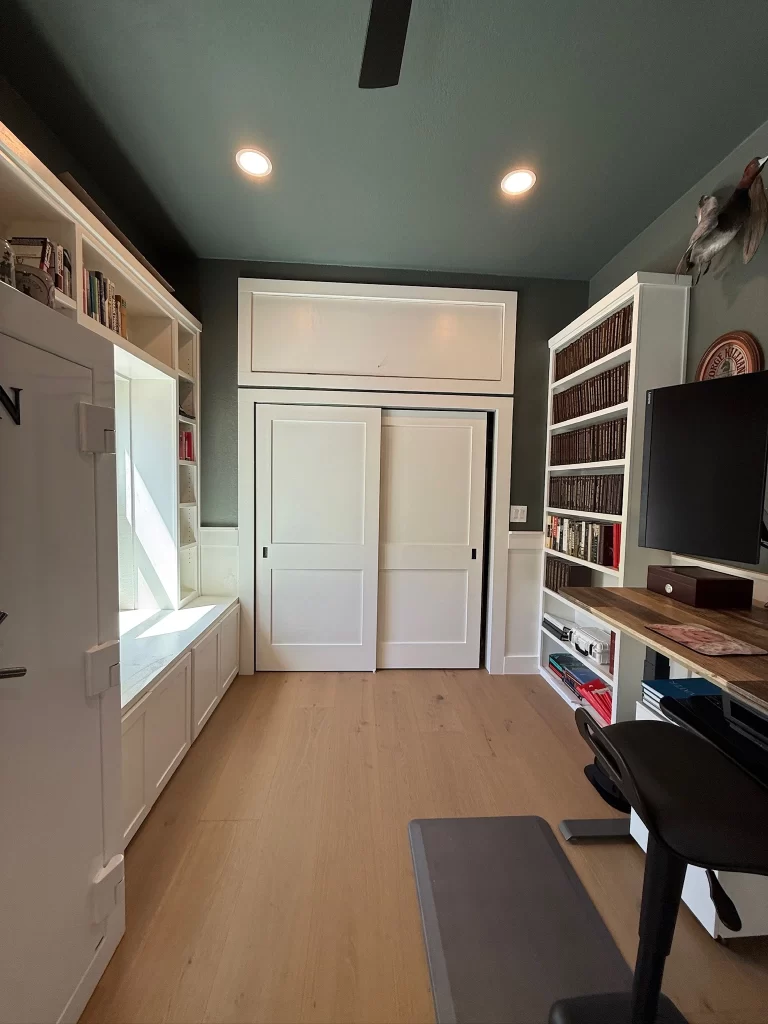 A spacious home office featuring a wooden desk with a black chair, built-in bookshelves filled with books, and light-colored wooden flooring. The walls are painted dark, with recessed lighting and a ceiling fan.