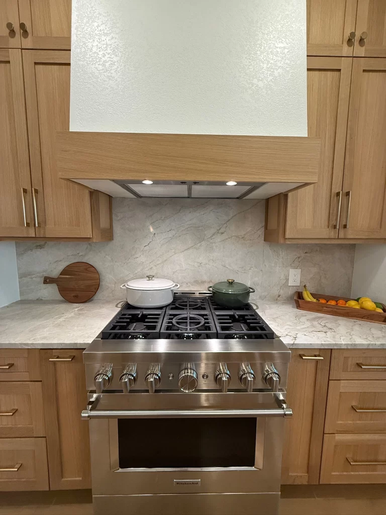 A modern kitchen featuring a stainless steel gas stove with knobs, a range hood above, and two cooking pots on the burners. The countertop is made of light-colored marble, and there are wooden cabinets with gold handles. To the right, a wooden tray holds various fruits.
