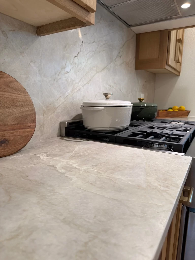 A modern kitchen countertop featuring light-colored stone, a wooden cutting board, and a white cooking pot on a gas stove, with wooden cabinets in the background.