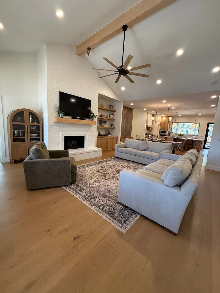 Cozy living room featuring a light-colored sofa, an armchair, a large rug, a white brick fireplace, a TV mounted on the wall, and a ceiling fan, with an open kitchen area visible in the background.