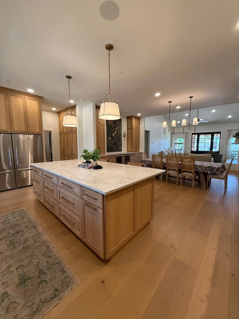 Modern kitchen interior featuring a large central island with a marble countertop and wooden cabinetry, illuminated by pendant lights. The background shows a dining area with a table and chairs, and a cozy atmosphere.