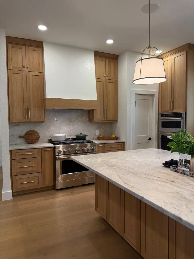 Modern kitchen featuring wooden cabinets, a marble island, stainless steel appliances, and a contemporary light fixture.