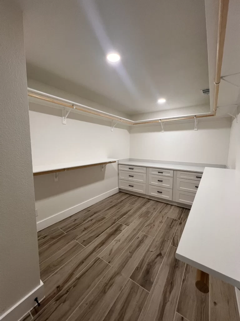 Spacious walk-in closet featuring built-in white drawers, shelving, and wooden flooring, illuminated by ceiling lights.