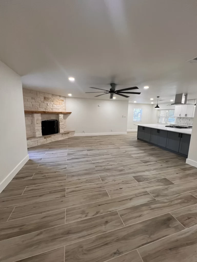 Spacious living room with wooden-style tile flooring, modern ceiling fan, and a stone fireplace featuring a wooden mantel, adjacent to an open kitchen with dark cabinetry.