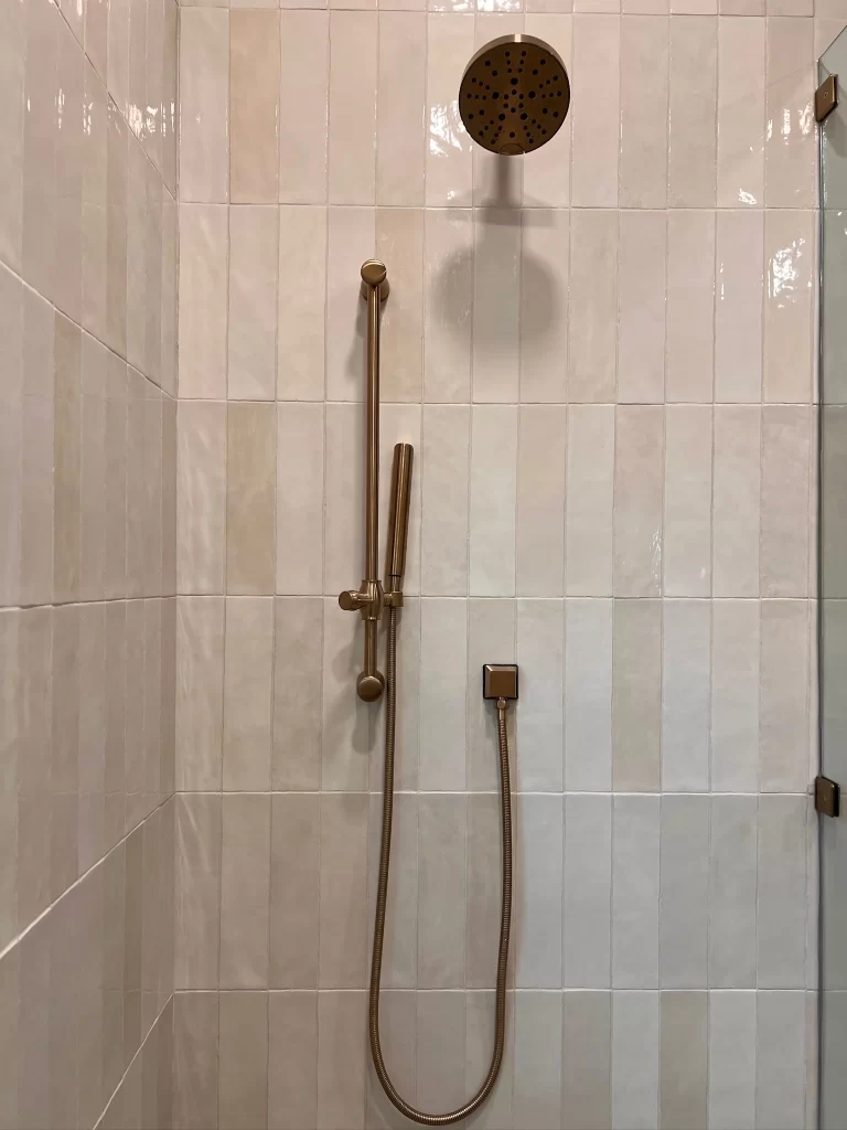A modern shower with a brass showerhead, featuring a handheld shower attachment, set against a backdrop of light-colored tiled walls.