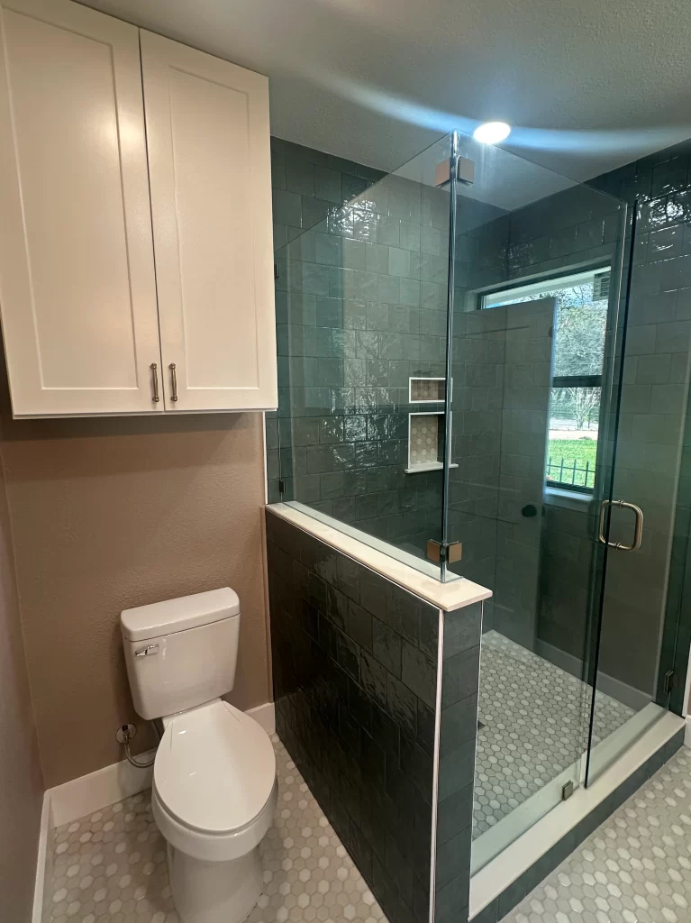A modern bathroom featuring a glass shower, a white toilet, and light-colored cabinetry against a tiled wall.