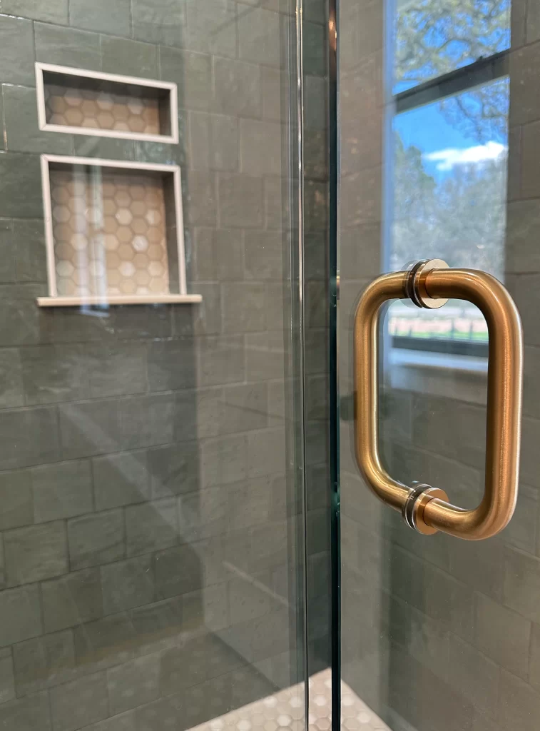 Close-up view of a modern shower door with a gold handle, featuring a glass panel and tiled wall with hexagonal patterns and a built-in shelf.