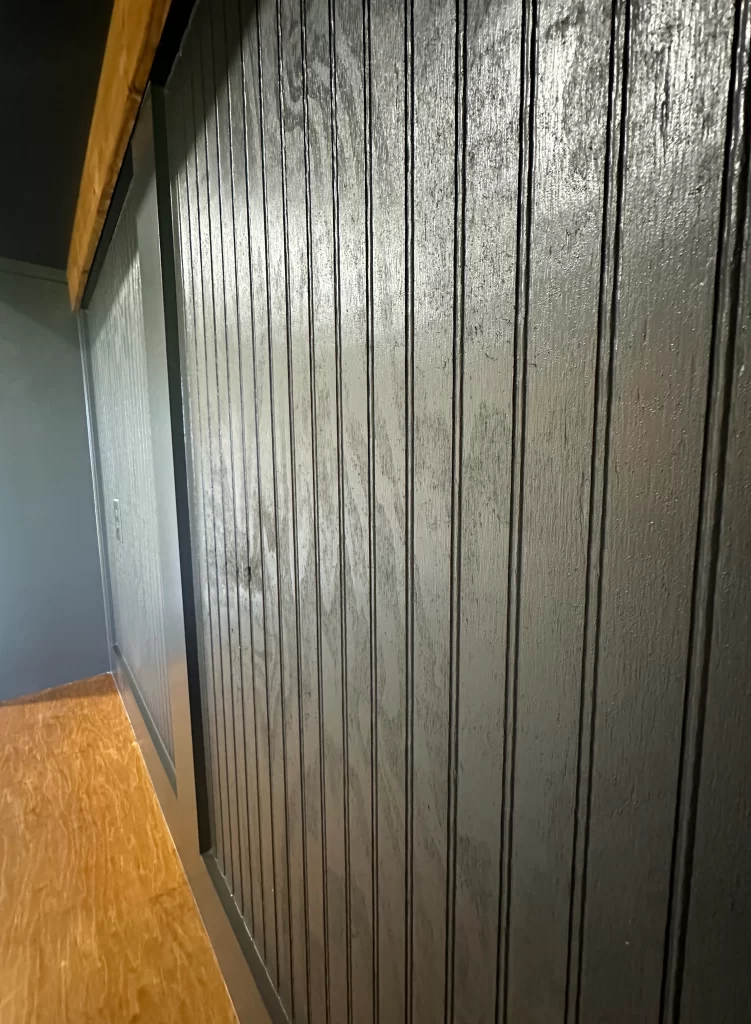 Close-up of a black wooden wall with vertical paneling and a part of a wooden floor visible.