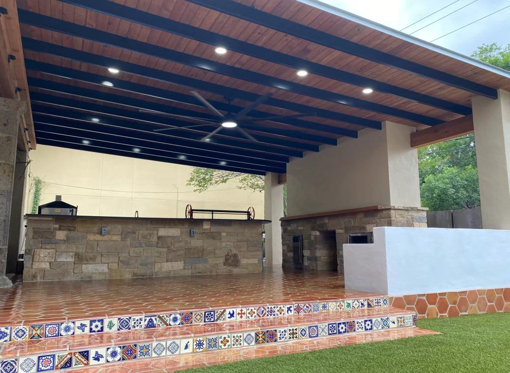 Outdoor kitchen and patio area featuring a large covered wooden structure with ceiling fans, stone walls, and decorative tiled steps.