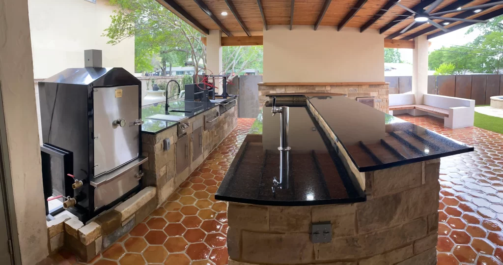 An outdoor kitchen featuring a large granite countertop, a smoker grill, and modern appliances, surrounded by a stone wall and a shaded area with a wooden ceiling.