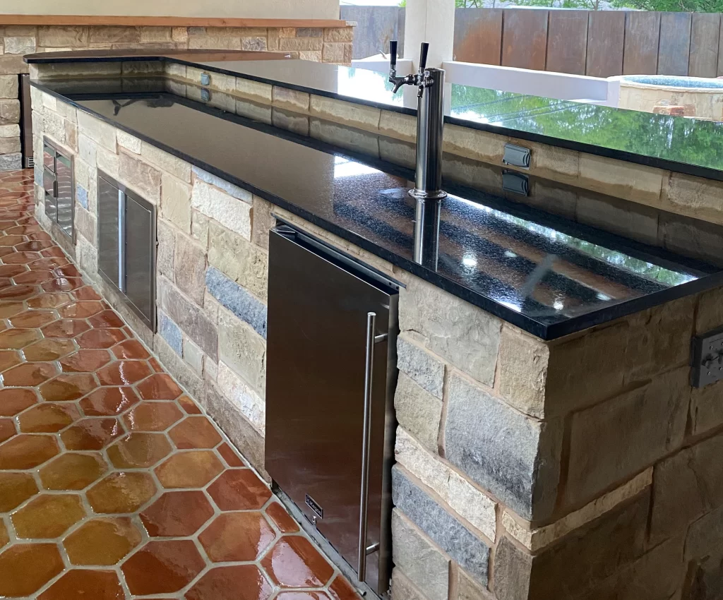An outdoor kitchen with a stone base and a black granite countertop, featuring a beer tap and stainless steel appliances below, on a patterned terracotta tile floor.